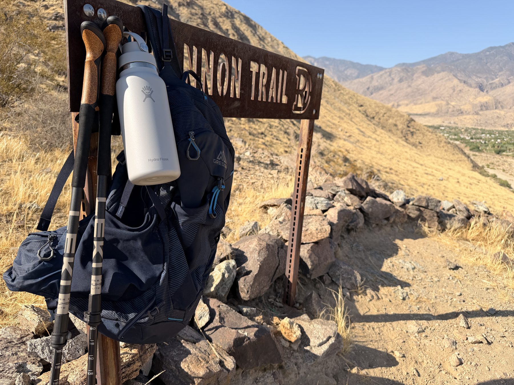 Gregory pack, Leki poles, and Hydro Flask bottle resting at a Palm Springs trail sign, Coachella Valley below